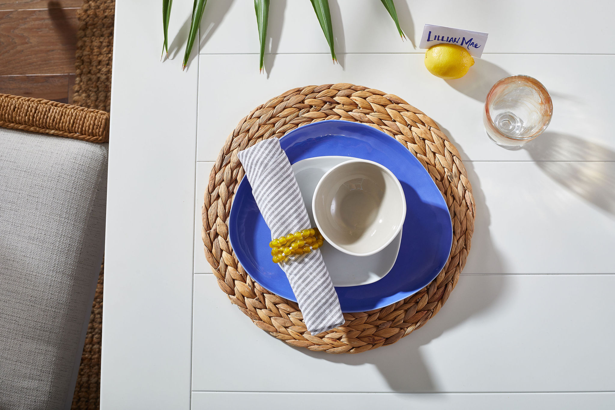 Place setting with a woven placemat, blue base plate, white bowls, a clear glass and a lemon with a nametag in it.